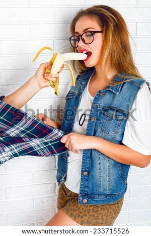 Young stylish sexy pretty hipster girl having fun, and surprised. and eating big banana from another hand. Urban white brick wall background.