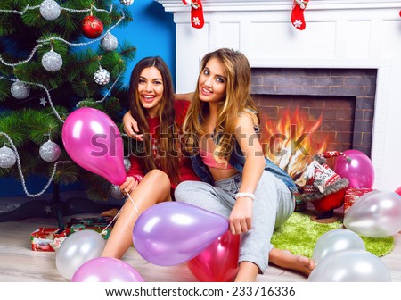 Winter lifestyle portrait of two sisters sitting at home near Christmas tree, holding balloons, ready for holidays party. Wearing bright make up and clothes. Best friends having fun.