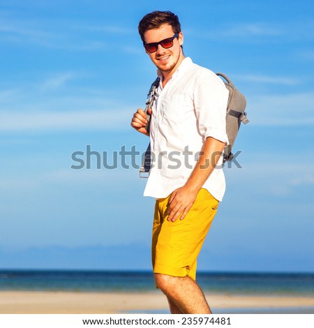 Outdoor fashion lifestyle image of young traveler handsome hipster man, wearing bright yellow shorts sunglasses, and holding his backpack, ready for adventures. Amazing view on beach and blue sky.