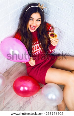 Young pretty girl having fun on birthday party, wearing party crown casual sweater, holding pink balloon and tasty small cake. Positive emotions, read for celebration.