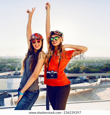 Outdoor lifestyle portrait of two crazy happy best friend girls screaming and put their hands to the air, enjoy amazing view from the roof at sunset, wearing trendy bright clothes caps and sunglasses.