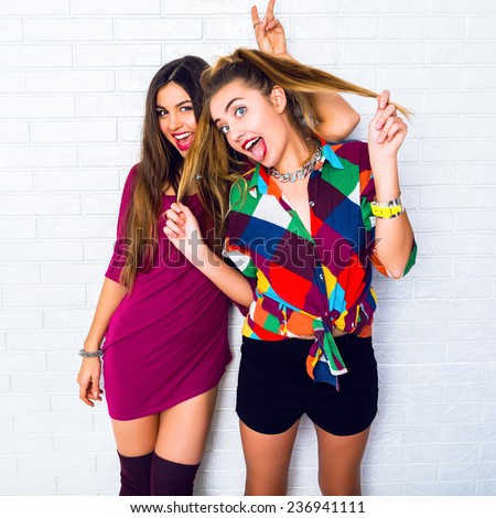 Lifestyle image of two young friend girls making crazy funny faces, wearing bright hipster clothes, urban white background. Showing tongue and screaming.