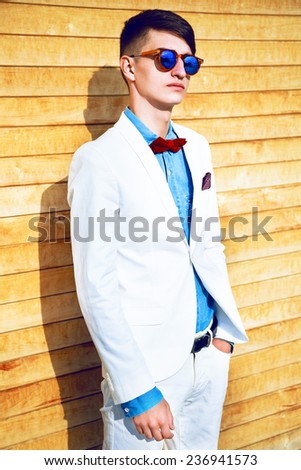 Fashion portrait of stylish man with hipster haircut, wearing trendy elegant white suit, denim shirt, bow tie and mirrored sunglasses, posing near wooden wall. Street style look.