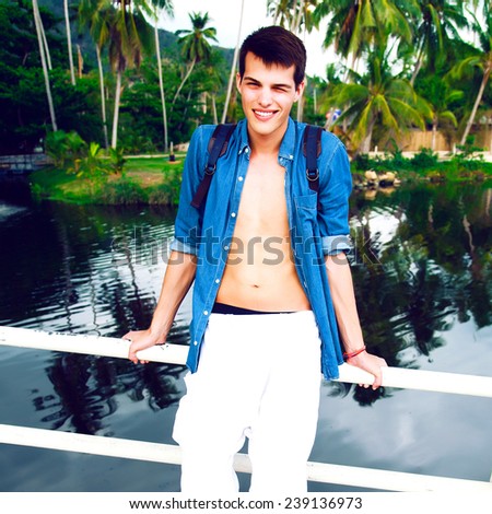 Outdoor fashion portrait of young handsome guy smiling and having fun near lake on exotic island, travel with backpack, wearing stylish denim shirt.