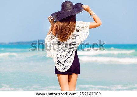 Outdoor summer portrait of young pretty woman looking to the ocean at tropical beach, enjoy her freedom and fresh air, wearing stylish hat and clothes.