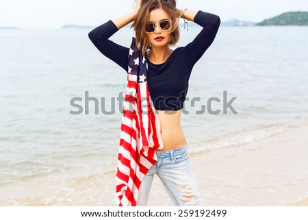 Outdoor fashion portrait of young sensual woman, wearing retro old school denim jeans crop top and sunglasses,  holding american flag, posing near sea at rainy day, romantic atmosphere, vintage style.