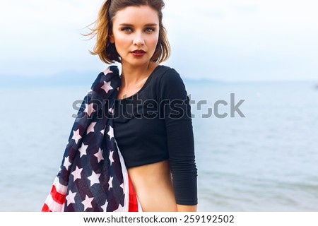 Young beautiful woman posing at sea side, holding american flag, wearing crop top and gothic bright make up. Rainy day, foggy weather.