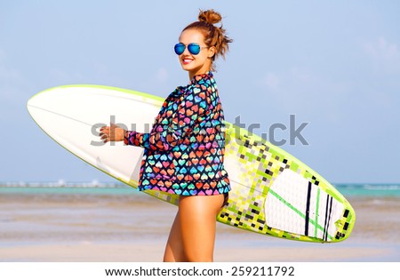 Outdoor summer portrait of sexy smiling woman running with surfer board near blue ocean,  wearing bright neon clothes and sunglasses, have fit tanned body and ginger hairs.