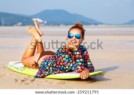Outdoor fashion lifestyle summer portrait of stunning sexy surfer woman, wearing bright outfit makeup and sunglasses. Laying on surfboard, amazing view on mountains and empty beach.