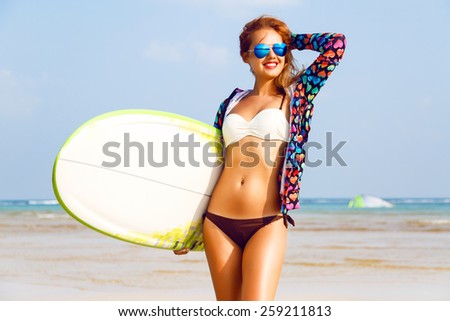 Outdoor fashion lifestyle summer portrait of stunning sexy surfer woman, wearing bright outfit makeup and sunglasses. Posing near clear blue ocean, enjoy her vacation.