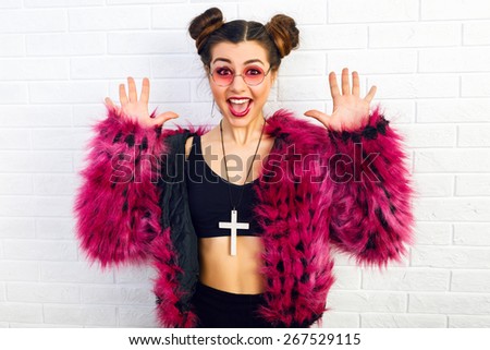 Indoor fashion portrait of stylish young woman smiling and  having fun alone, wearing trendy hot pink fake fur coat and round sunglasses, eighties style. Urban white wall background.