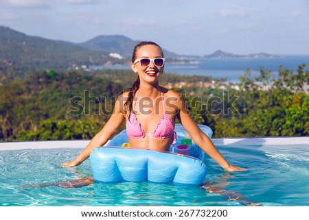 Young sexy woman in bright pink bikini and vintage sunglasses having fun and swimming in air mattress at pool party, on luxury villa, amazing view on tropical island and ocean.