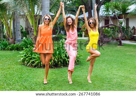 Outdoor lifestyle fashion portrait of the pretty girls friends having fun on vacation, wearing stylish bright neon dresses and sunglasses. Jumping and dancing at tropical garden.