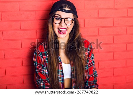 Young pretty seductive hipster woman having fun and showing long tongue and winked, wearing plaid shirt and clear glasses, long hairs and bright make up. Red urban wall background.