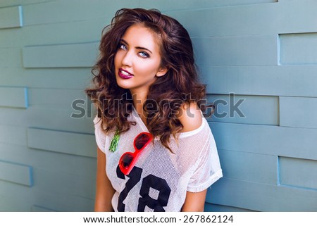 Close up bright fashion portrait of pretty young teen hipster woman with bright make up and stunning curled fluffy brunette hairs, wearing sportive outfit and neon sunglasses, urban background.