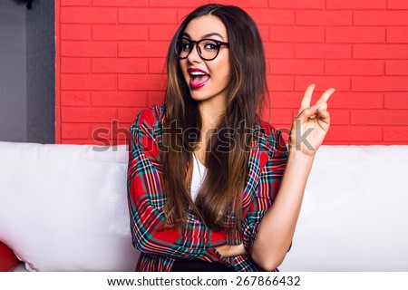 Close up indoor portrait of young pretty hipster girl with long brunette hairs and bright make up, showing yo science and laughing, red urban wall background.