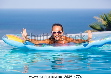 Outdoor lifestyle portrait of stunning young woman having fun at infinity pool with amazing view on tropical island, wearing bright bikini and sunglasses, swimming on air mattress.