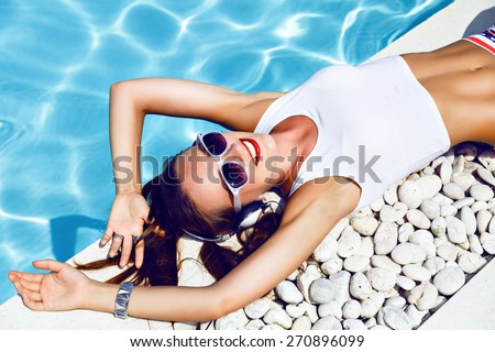 Summer fashion portrait of young sexy dj girl laying near pool, wearing sexy mini shorts with stars vintage sunglasses, cute hairstyle and bright make up, listening music on headphones.