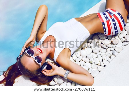 Summer fashion portrait of young sexy dj girl laying near pool, wearing sexy mini shorts with stars vintage sunglasses, cute hairstyle and bright make up, listening music on headphones.