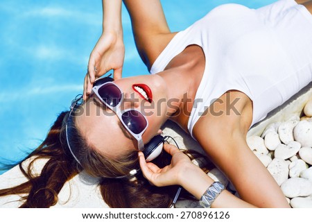 Close up fashion portrait of cute smiling young girl, listening and enjoy music on headphones, laying on her vacation her pool, wearing ponytails sunglasses and bright make up.