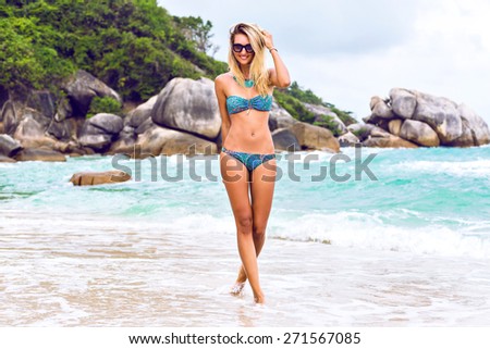 Summer lifestyle portrait go young sexy blonde woman with fit tanned body, wearing stylish bikini and sunglasses, having fun on island beach. walking alone and thinking on someone. romantic mood.