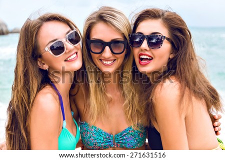 Lifestyle close up fashion portrait of three pretty girls best friends having fun smiling and posing on camera at the beach near ocean, wearing stylish bright bikini and sunglasses.