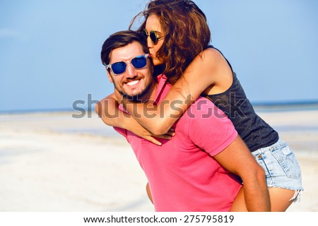Young happy couple in love having fun and going crazy at the beach at nice sunny summer day, young woman jumping on her boyfriend, positive emotions.