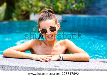 Summer trendy close up fashion portrait of sexy tanned woman relaxed and swimming at pool. wearing bright bikini and sunglasses, smiling and looking on camera. positive mood.