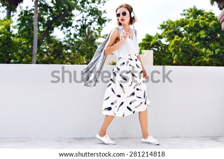 Young stylish hipster woman walking alone at nice hot summer day, listening music in her earphones, wearing vintage trendy outfit and sunglasses.