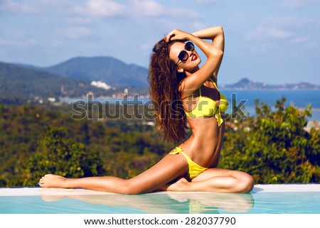 Young sexy woman siting and relaxed near pool at summer holiday nice hot  day, wearing yellow bikini and stylish sunglasses in tropical island. Sexy perfect fit body lady.