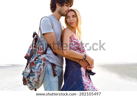 Outdoor lifestyle portrait of amazing pretty hipster young couple in love posing on the beach. Stylish man and woman hugs and spend great time together. floral dress backpack and denim.
