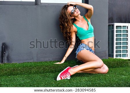 Girl with a slim figure in a bright top, denim shorts and sunglasses,long hair, tanned body sitting on the grass near the wall.Bright colors have a good mood. Urban style.