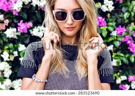 Young gorgeous stunning blonde young woman , wearing stylish outfit, trendy sparkled crop top and sunglasses, posing in floral garden background, beautiful summer day.