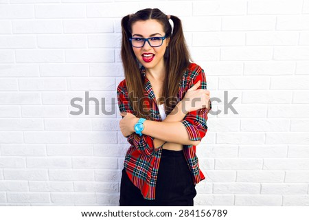 Young funny cheeky teen girl, making funny angry face, and screaming, bright make up, long butte hairs, hipster glasses and plaid shirt, going crazy alone. White urban brick wall background.
