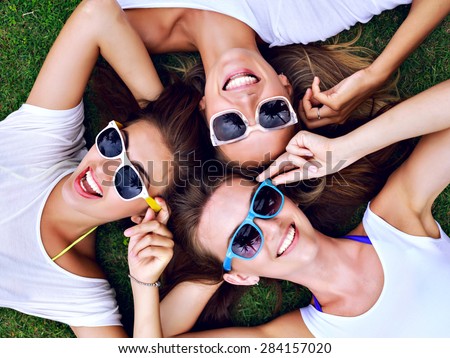 Summer lifestyle portrait of three hipster woman laying on the grass enjoy nice day, wearing white simple tops and bright sunglasses. Best friends girls having fun, joy, playful mood.