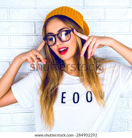 Funny emotions. Beautiful young blond haired woman in glasses and a yellow hat, making face, standing up against a light brick wall background