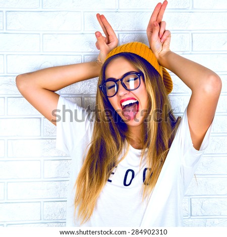 Young girl doing emotion, hands raised up in the form of rabbit ears . Dressed in a white shirt, yellow hat, glasses and bright lips, trendy clothes.