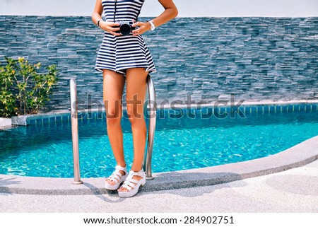 Cropped image of young woman in high heeled shoes standing on the edge of the pool wearing a dress with a good figure. holding vintage camera, summer portrait of photographer hipster girl.