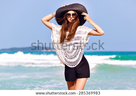 Summer fashion portrait of beautiful woman enjoy windy sunny day near ocean, vacation style. Young stylish girl wearing black romper vintage hat and big sunglasses, bright colors.