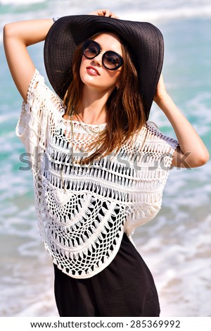 Summer fashion portrait of beautiful woman enjoy windy sunny day near ocean, vacation style. Young stylish girl wearing black romper vintage hat and big sunglasses, bright colors.