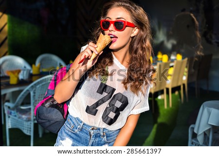 Charming beauty.Beautiful young woman eating a big sweet ice cream in red sunglasses, shorts, sports bag on his shoulder, standing   outside in summer.