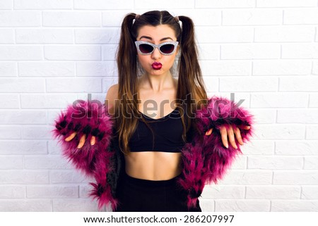 Personal style. Cute girl with glasses and funky wear purple  faux fur coat  and a tied of hair raised her hands on a white background.  Crazy disco style.