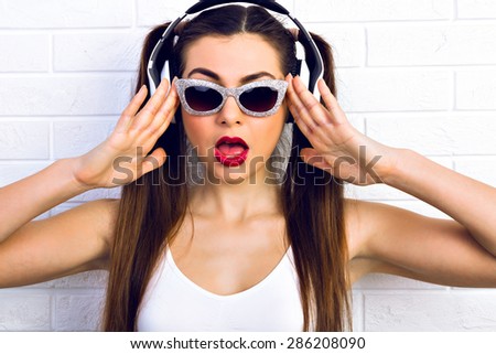 Wow! Excited young woman surprised looking at the camera wearing sunglasses, headphones, listening to music on a white background. Surprised emotions. ready for party.