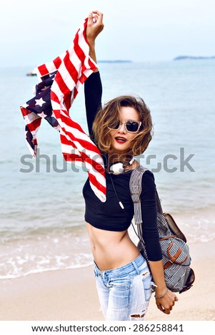 Young pretty woman having fun on the beach, jumping dancing and screaming, holding american flag, spending time near ocean. travel with backpack.