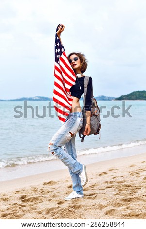 Summer fashion portrait of stunning fit sexy woman, wearing denim and crop top, holding american flag, spend time at the beach at rainy day, jumping and having  fun.