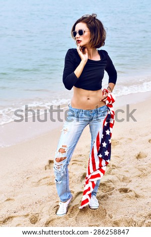 Fashion sensual portrait of beautiful sad woman posing on the island beach at windy rainy day, wearing old school outfit, have fit sexy bode, holding american flag in her hands.