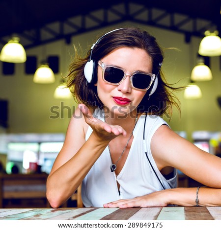 Fashion bright portrait of stunning happy woman sending air kiss, having fun, listening music on her headphones, toned colors.