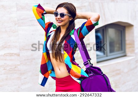 Bright summer trendy portrait of young fit girl posing on the street, wearing vivid shirt crop top and leg gins holding big sportive bag, long hairs and make up. walking alone after workout .