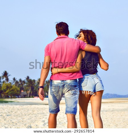 Summer fashion image of stylish hipster couple posing at lonely exotic beautiful beach, hugs and kisses, joy , enjoy their vacation, bright trendy clothes, sunny colors, positive mood.