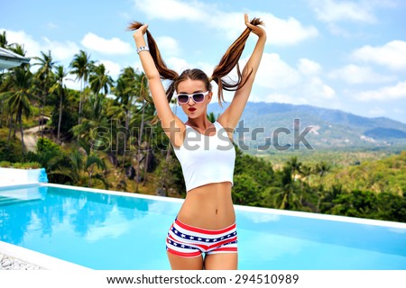 Summer bright portrait of sexy hipster girl having fun at pool party, holding her ponytails and flirting, joy, vacation, tropical island.